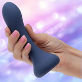 A hand with pink-painted nails holds the CalExotics Deep Curve Vibrating Probe, a dark blue silicone massager, against a softly blurred purple and pink background.